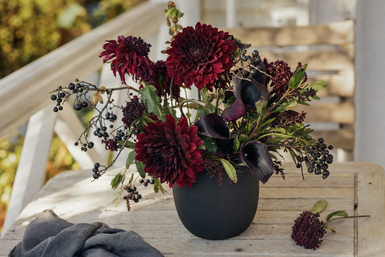 A dark burgundy and plum bouquet with calla lilies and berried branches in a charcoal vase in bright daylight